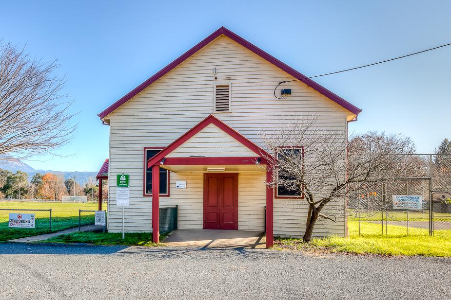 The exterior of Porepunkah Hall, a single-story building clad in cream weatherboard with red trim around the roofline and entryway.