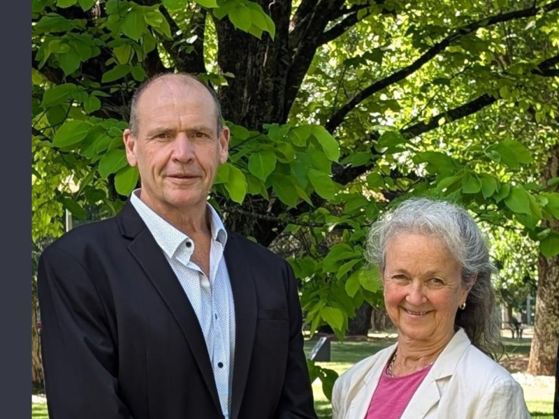 A tall white man in a white shirt and black blazer standing to the left of a white woman with white hair, pink shirt, and white blazer.