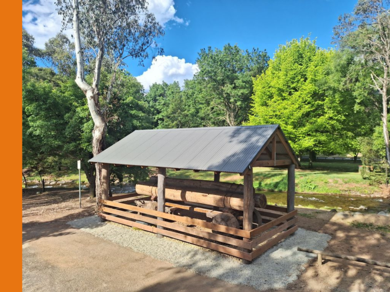 An open shed, constructed of timber with a tin roof, covers a historic log wagon. The shed is located next to a creek.