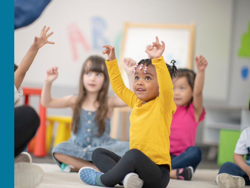 Kindergarten students sit in a classroom with their hands in the air, all looking to the left at a person out-of-frame. The child in focus has brown skin and is wearing a yellow long-sleeved shirt, black pants, and black hair braided and secured with beads.