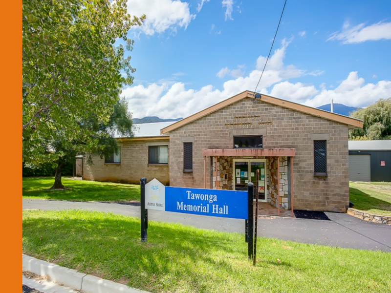 The exterior of Tawonga Memorial Hall, a single-story brown brick building with a peaked roof and inset entryway. The glass entry doors are adorned with colourful posters.