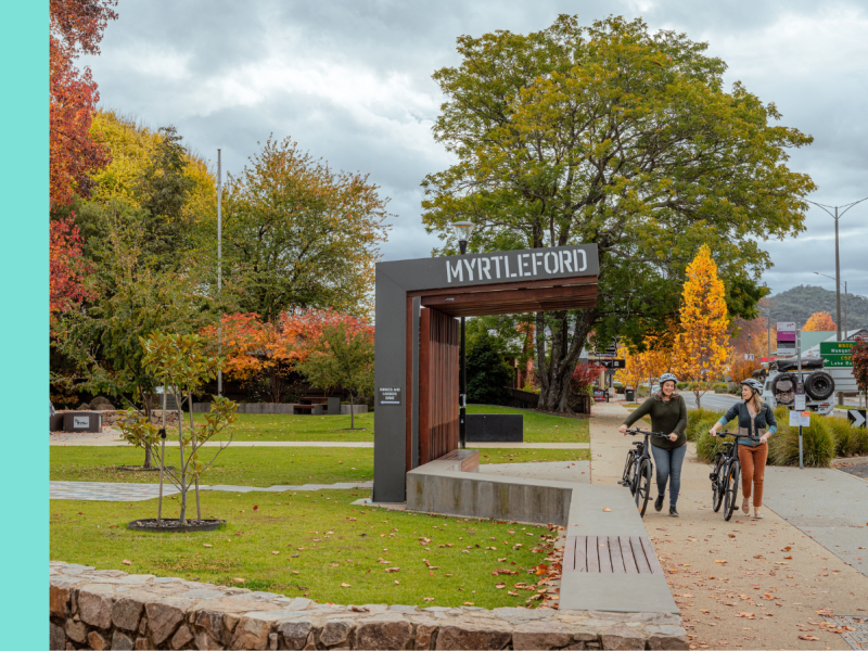  The image shows two people walking their bicycles near the entrance sign for Myrtleford, a town with a mix of greenery and autumn-colored trees in the background. The sign is modern, with large white lettering, and stands in a landscaped area with paved pathways and a stone wall. The atmosphere is relaxed and inviting, with a cloudy sky above and vibrant foliage surrounding the scene.