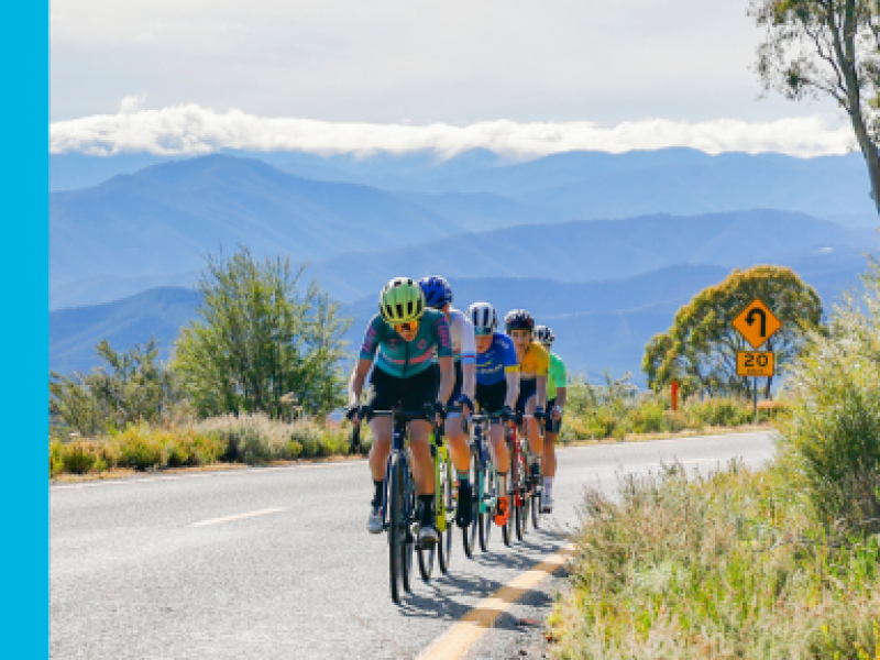  A group of cyclists rides along a winding road with mountainous scenery in the background. The cyclists wear colorful jerseys and helmets as they ride in a single file line, with a yellow road sign indicating a curve ahead. The landscape features green vegetation, and the distant mountains are partially covered by clouds, creating a scenic outdoor setting.