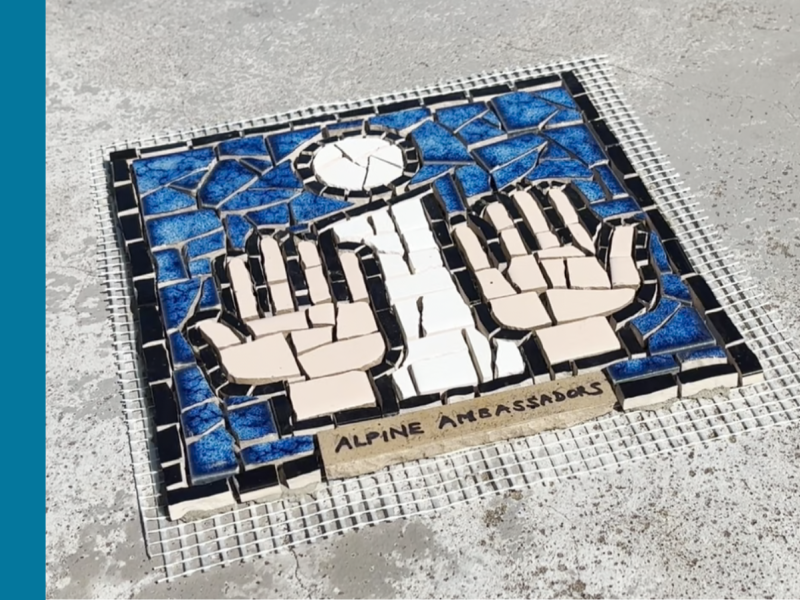 A mosaic tile on a concrete surface depicting a white 'i' with two hands on either side. The words Alpine Ambassadors is written on a beige tile underneath the i and the hands.