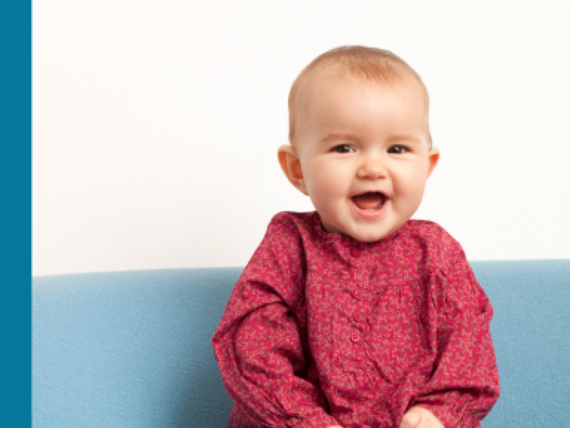 A white baby smiles at the camera, wearing a red, long-sleeved top.