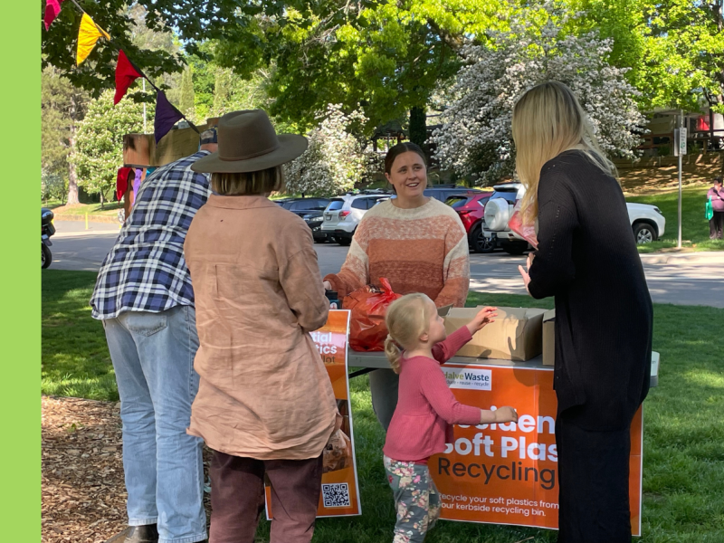 A pop-up stall in a grassy area with a carpark immediately behind. A young woman is smiling at a woman, who is facing her. An orange poster attached to a table reads Soft Plastics Recycling.