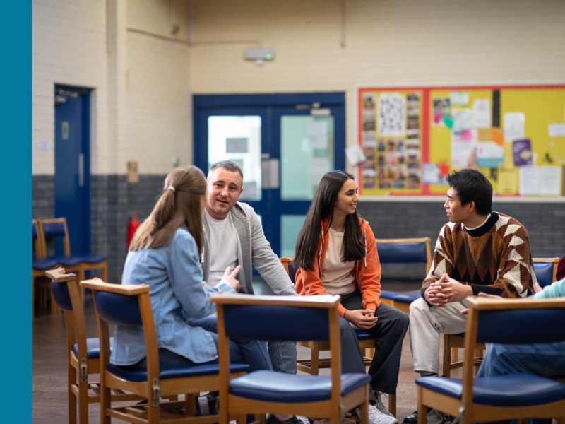 Four people sit on chairs, which are arranged in a circle. The man and woman on the left are talking to each other, while the young woman and young man on the right are talking to each other.
