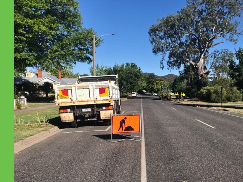 Truck parked on side of road with orange works ahead sign. Outdoor crew in the background cleaning up a fallen tree on the road.
