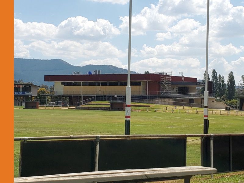 The Ablett Pavilion in Myrtleford under construction with large boards up over the windows and doors, and football goal posts in front with the Pavilion in the background.