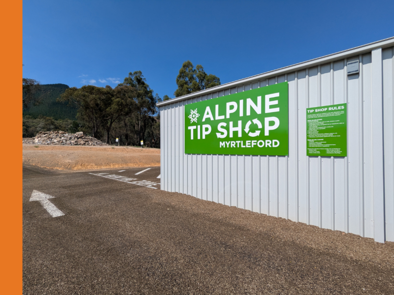 The exterior of a shed, cladded in white Colourbond. There is a green sign with the words Alpine Tip Shop Myrtleford on the side of the shed. 