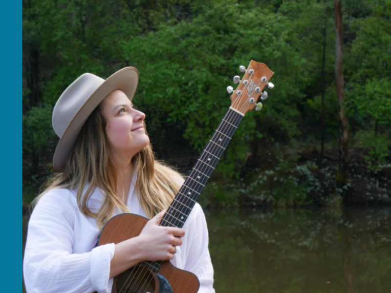 A young white woman holding a guitar with one hand while looking up to the sky.