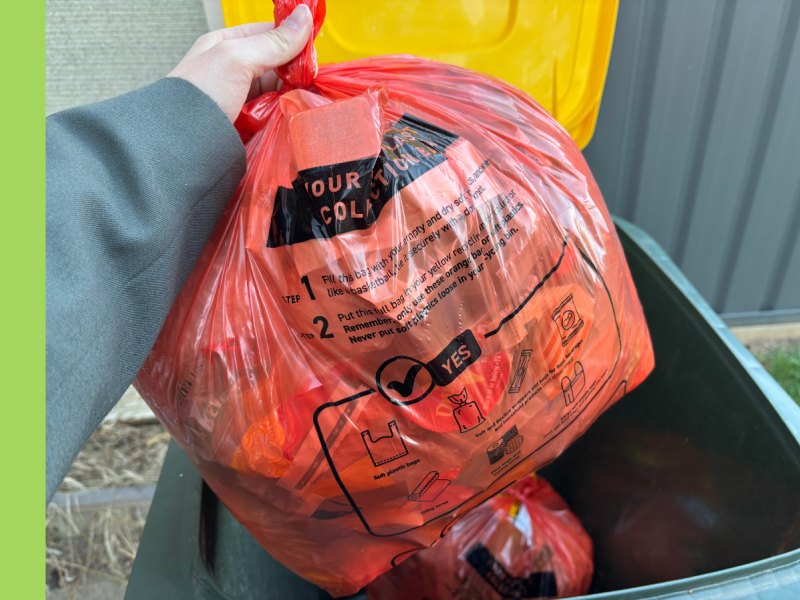 An orange bag filled with soft plastics held above a yellow-lidded kerbside recycling bin.