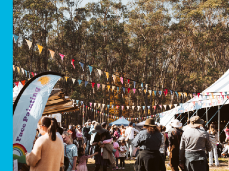 A loose crowd between market stalls on the left and a large marquee on the right, with colourful bunting fluttering above their heads.