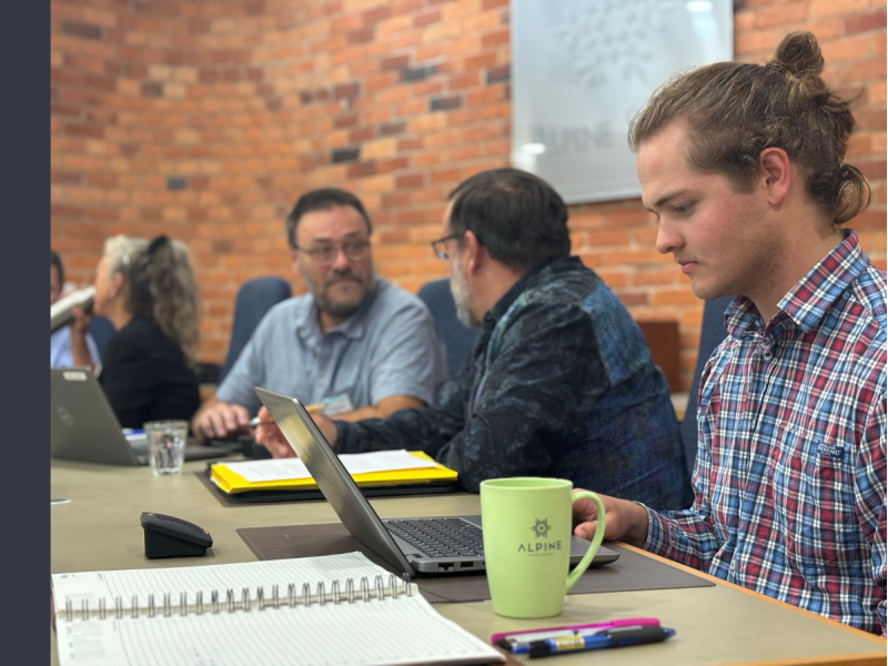 A young man in a collared red and blue plaid shirt is sitting at a table, looking down at a laptop in a room with red brickwork. There are two men, sitting at the same table, having a conversation behind him.