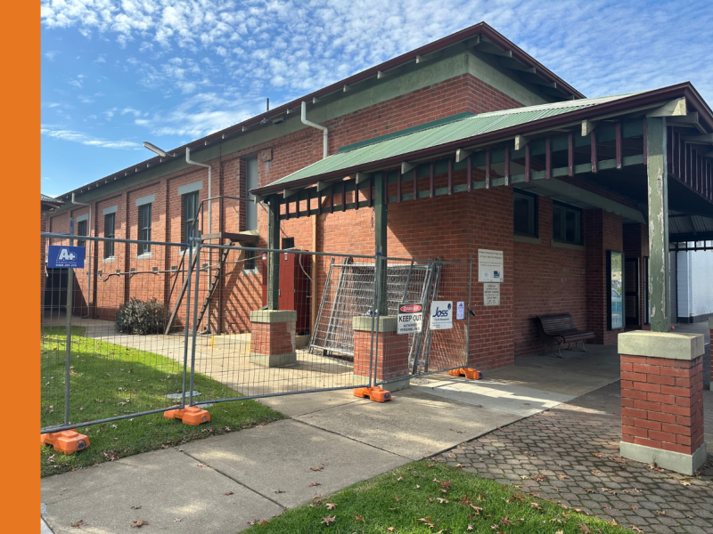 The exterior of Myrtleford Memorial Hall, a red brick, single-story building with a green roof and trim. There is temporary fencing erected to the left of the Hall.