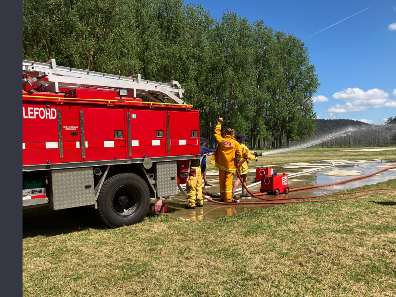 A CFA firefighter helps a young child spray water from a hose. They are standing behind a red firetruck in an open grassy field with green trees to their left.