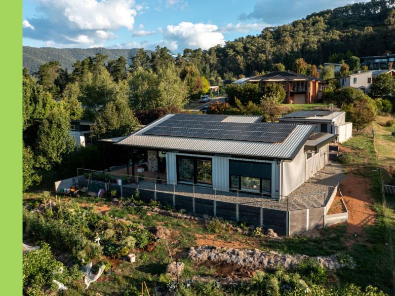 A single-story house on a hill with solar panels on the roof and a retaining wall above a native Australian garden. 