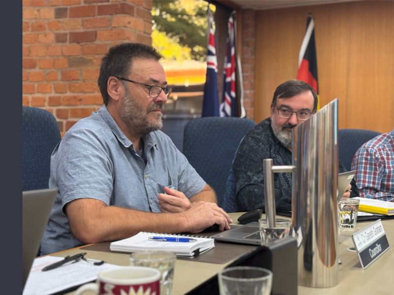 Two men sit at a desk in Council Chambers. The man closest to the camera has a blue short-sleeved shirt with a notebook and pen on the desk in front of him.