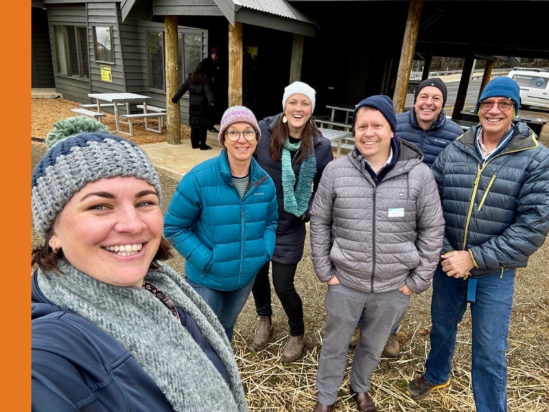 A woman wearing a blue and grey beanie and a grey scarf takes a selfie with two women and three men, who are all dressed for snow conditions.