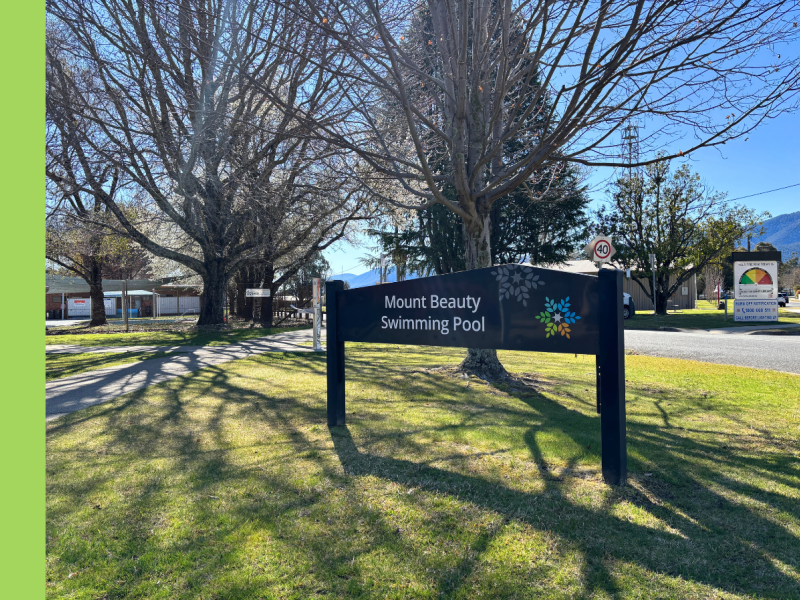 A blue facility sign installed in a park with the words Mount Beauty Swimming Pool printed on it. Behind the signs there are winter trees with no leaves on them.