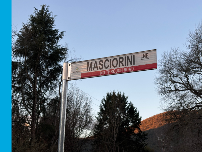 A street sign with the name Masciorini Lane printed on it in black text on a white background, as well as a logo of a snowflake with the word Alpine printed underneath the snowflake and the words No Through Road printed under the street name in white text on a red background.
