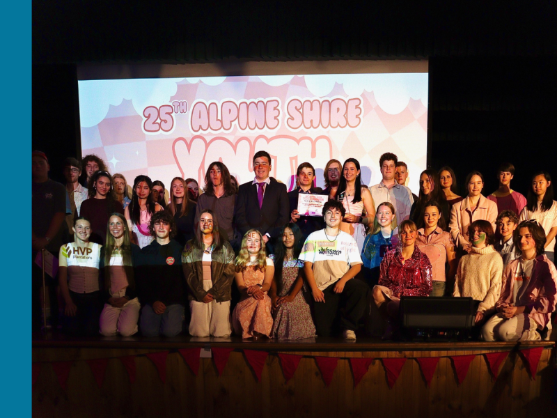 35 young people pose for a photo together on a stage with a presentation behind them that reads 25th Alpine Shire Youth Awards.