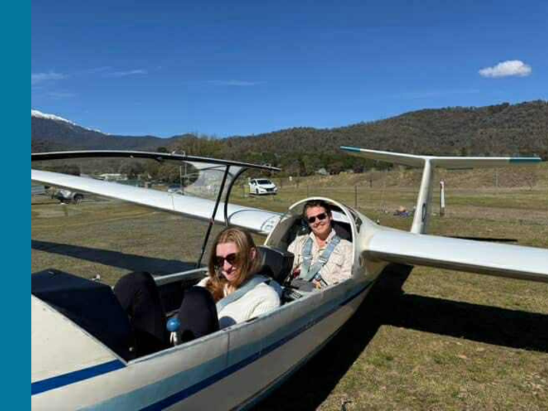 Two young people sit in a glider plane. They both have sunglasses on, with the person in the back smiling at the camera.