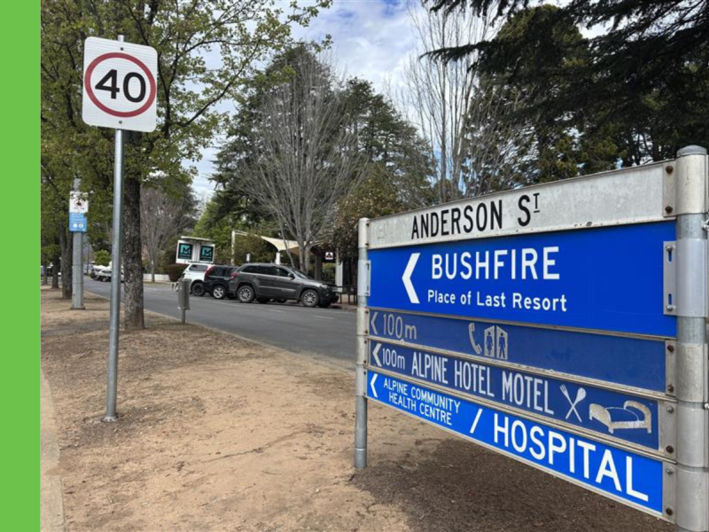 A cluster of road signs located on a median street that read from top to bottom: Anderson St, Bushfire Place of Last Resort, 100m to (phone symbol and toilet symbol), 100m Alpine Hotel Motel, and Alpine Community Health Centre / Hospital.