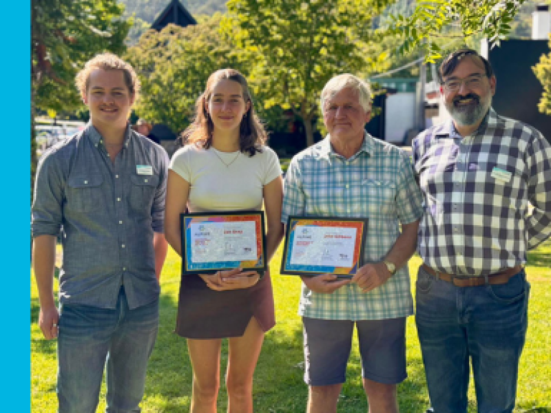 A young man, young woman, older man, and middle-aged man stand next to each other in a row from left to right. The young woman and older man are holding certificates. Everyone is looking and smiling at the camera.