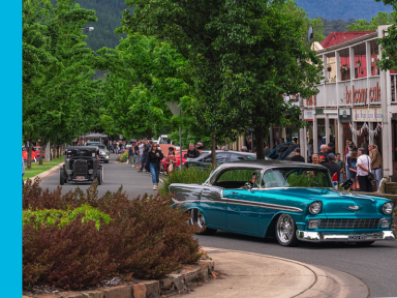 A classic car turquoise and silver in colour drives around a round about in Bright, with a crowd of people walking down the footpath in the background.