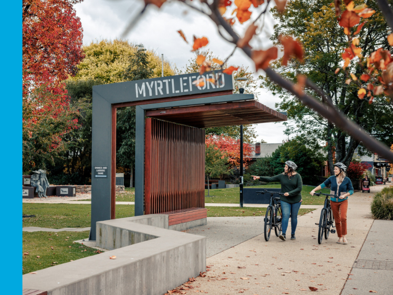 Two women walk their bicycles next to a wooden shelter with the word Myrtleford on it.