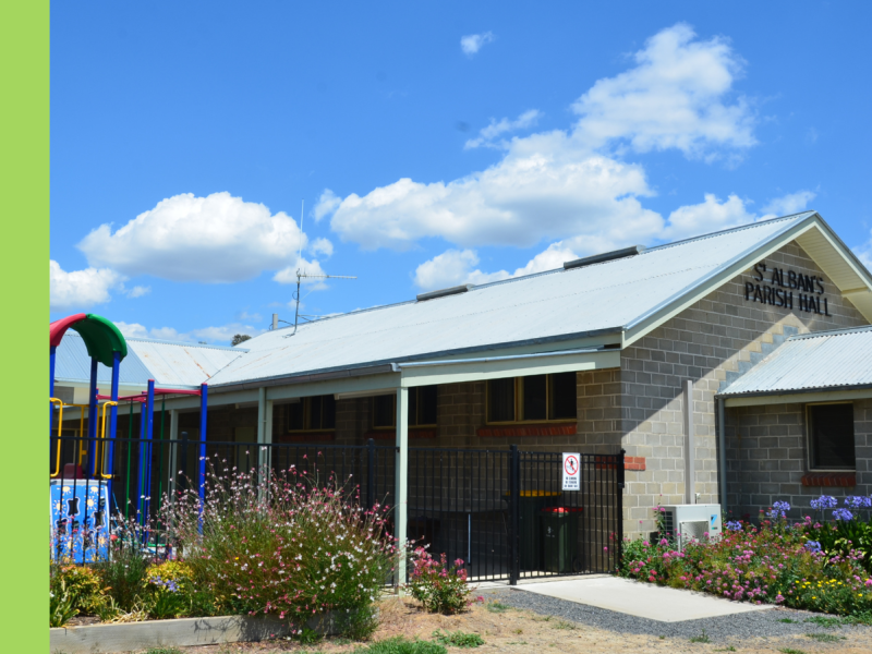 The exterior of a hall with grey brick and wildflowers growing along a path to a side gate. There is a playground to the left of the hall.