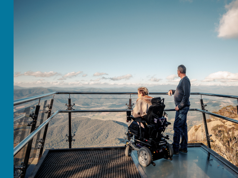 A woman in a motorised wheelchair and a man standing next to her look out over mountains from a lookout platform.