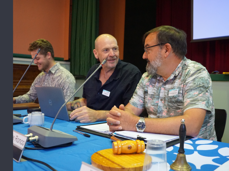 Three men sit at a bench with microphones in front of them. They are all smiling.