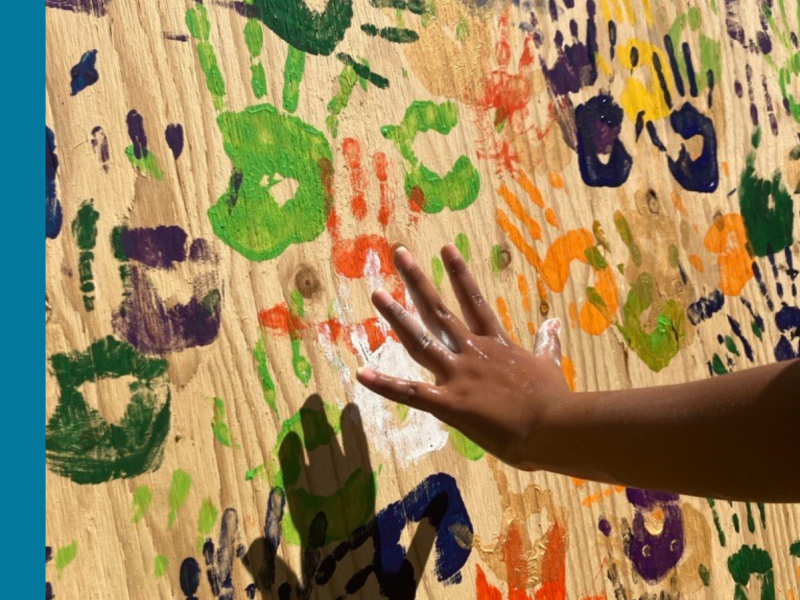 A left hand belonging to a young person reaches out towards a large poster of brown paper already covered in colourful handprints with the intention of adding another print to the wall.