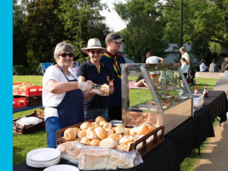 Two women, one wearing a broad brimmed hat, stand next to a young male in a baseball cap, in front of a tray of bread rolls. The first woman holds a sausage in tongs and is placing it in a bread roll.