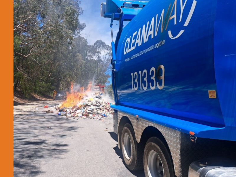 An image of the back half of a blue recycling truck with 'Cleanaway' written on the side. A pile of burning rubbish is located on the road behind the truck. Trees are in the background, suggesting a remote or bush location.