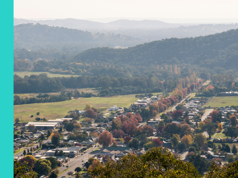 An aerial view of the main street in Myrtleford, showing autumn trees and rooftops, with mountain scenery in the background.