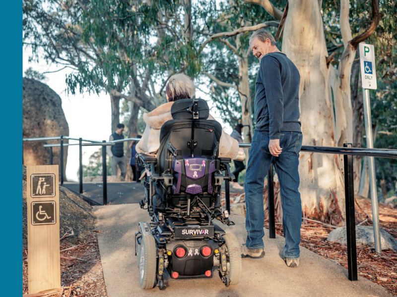 Woman in electric wheelchair looking up at man walking beside her as they travel up a ramp towards a lookout.