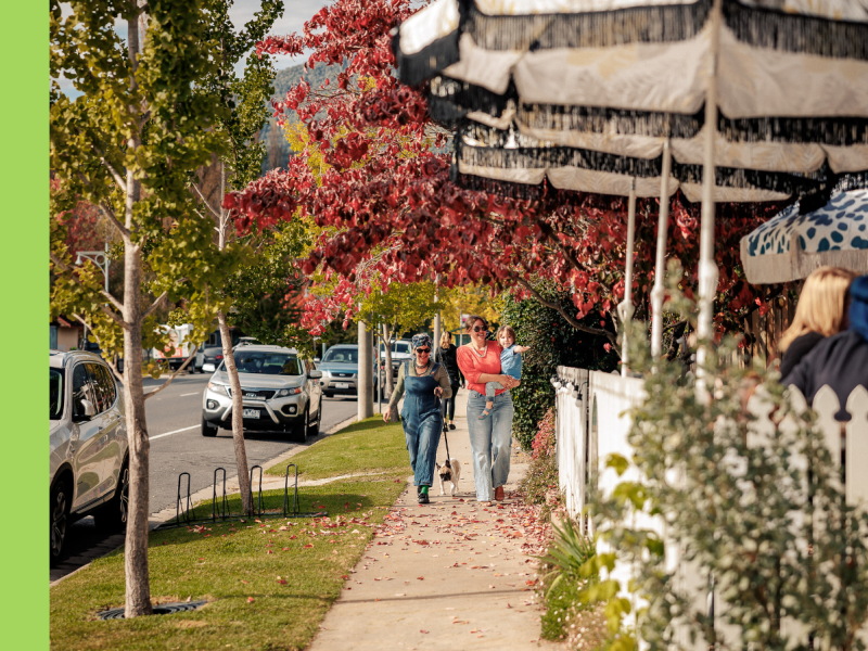 Two women walk down a street with shopfronts to the right and road to the left.
