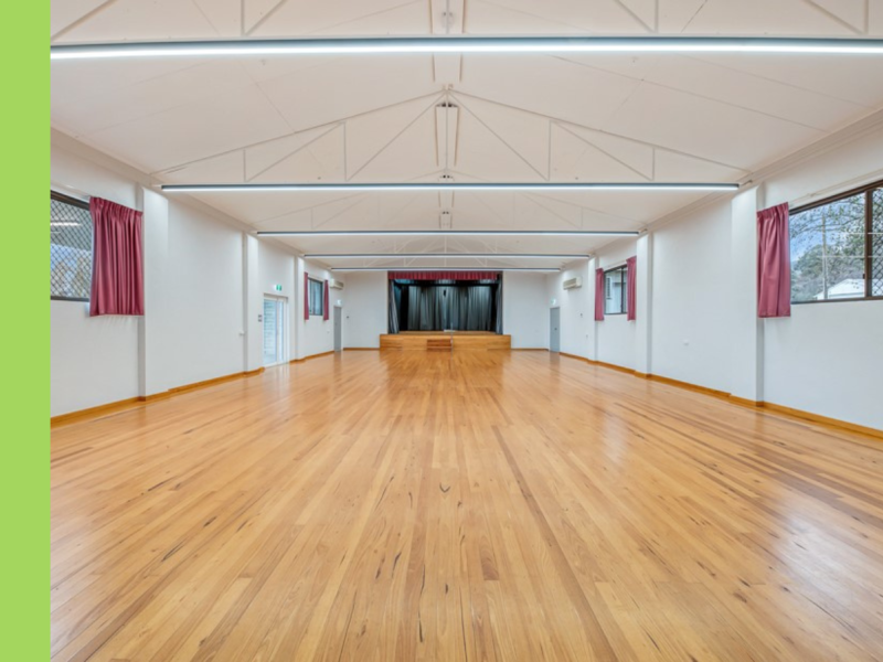 A hall with a stage at the far end, blonde wooden floors, and red curtains on the windows.