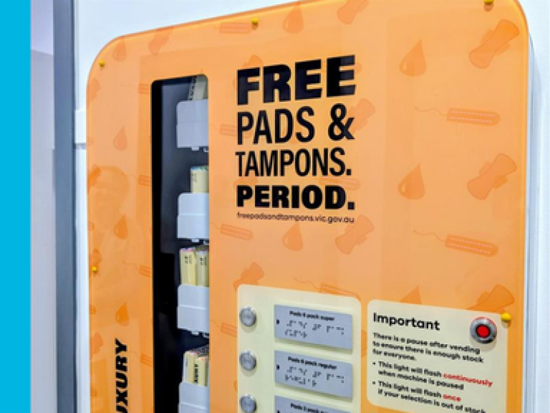 A close up of an orange vending machine with words printed in black across it that read FREE PDS & TAMPONS. PERIOD. Selection buttons and instructions for the vending machine are below.
