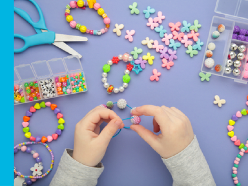 A child's hands thread a bead onto a half made bracelet, with the table in front of them scattered with colour beads of various shapes and sizes, along with a pair of blue scissors.