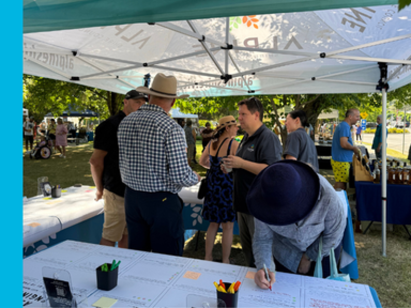 A group of five people standing under a marquee deep in conversation. In the foreground, a table is covered with paper with boxes and areas for feedback. A woman in a navy blue hat is leaning over the table writing something.