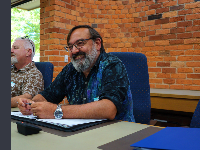 Two men sit at a desk in blue chairs in front of a red brick wall. The man closest to the camera has black hair and a white beard, and is wearing a short-sleeved blue shirt.