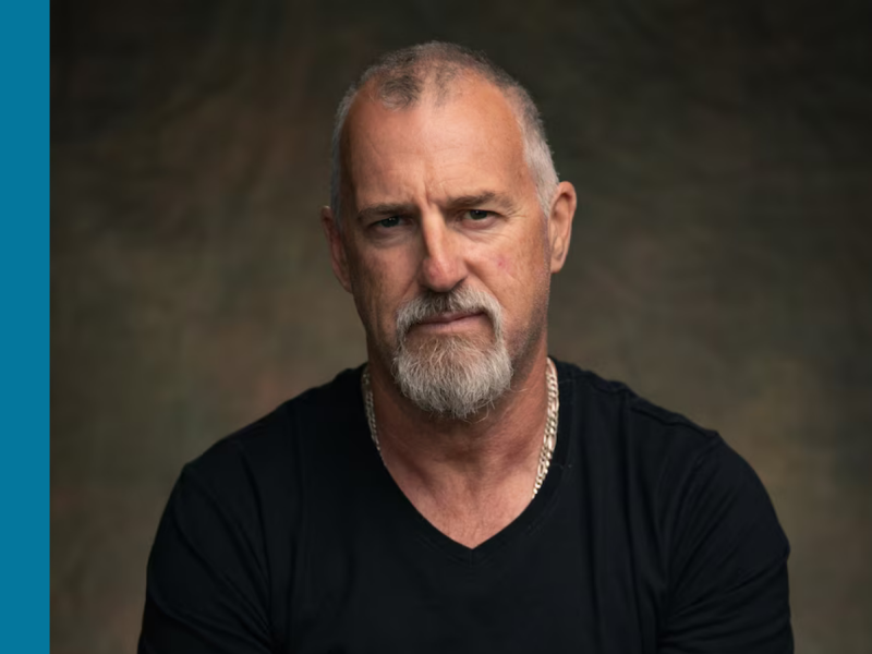 A middle-aged white man wearing a black t-shirt looks solemnly at the camera.