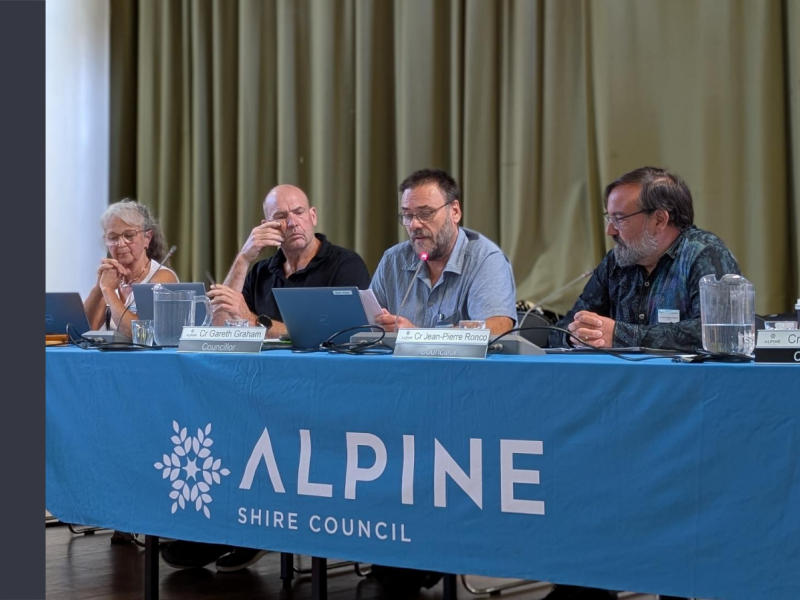 Four people sit at a desk that is covered with a blue tablecloth with Alpine Shire Council printed on it.
