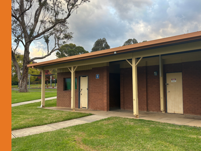 A red brick toilet building with cream doors and a beautiful gum tree in the background.
