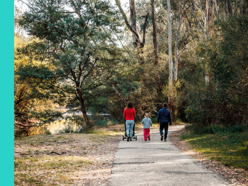 Three people, including a woman, a child, and a man, walking along a footpath. The woman is pushing a pram. We can only see their backs. There is lots of greenery in the background, suggesting a bush setting.