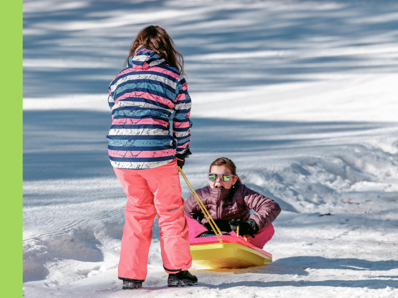 A young girl in a striped ski jacket and pink ski pants has her back to the camera, pulling another young girl wearing sunglasses and a purple ski jacket up a snowy slope on a yellow toboggan.